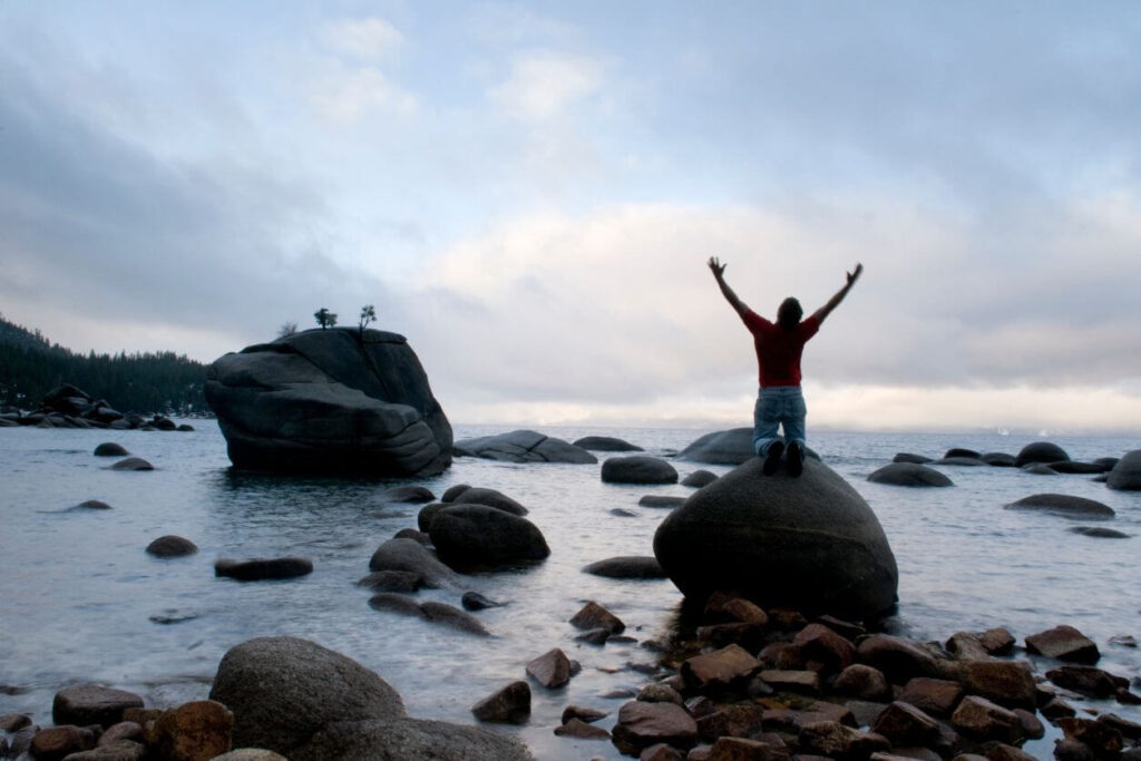 person on a shoreline kneeling on a rock with their arms stretched wide